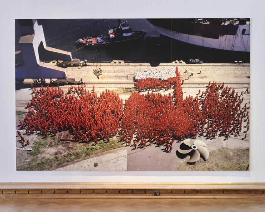 wielkoformatowe zdjecie wiszace na sciane w galerii sztuki wspolczesnej przedstawiajace ludzi ktorzy po tym jak byli ustawieni w kształt czerwonego napisu solidarność powoli się rozchodzą w różnych kierunkach, zdjecie jest z perspektywy lotu ptaka