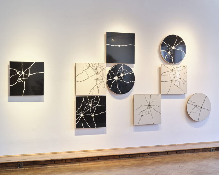 praca artystyczna przedstawiająca 3 kwadratowe i 2 okrągłe czarne oraz 3 kwadratowe i jedną okrągłą szarą płytkę ceramiczną, na ktróych widać ślady po strzałach kul