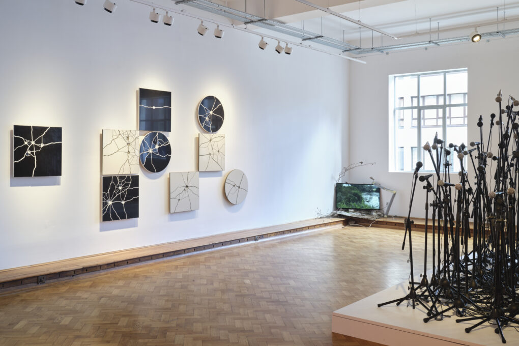 widok wystawy na której widać płytki ceramiczne w dwóch kolorach i kształtach ze śladami strzałów, ekran przy podłodze z projekcją wideo oraz podest na którym ustawiono wiele mikrofonów