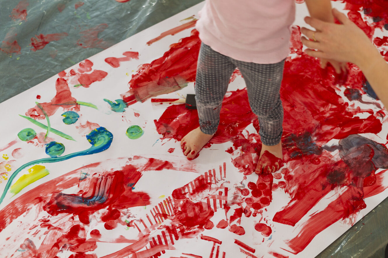 dziecko stojące na dużej kartce papieru malując stopami, kartka jest w plamach wielu kolorów ale dominuje tam czerwień