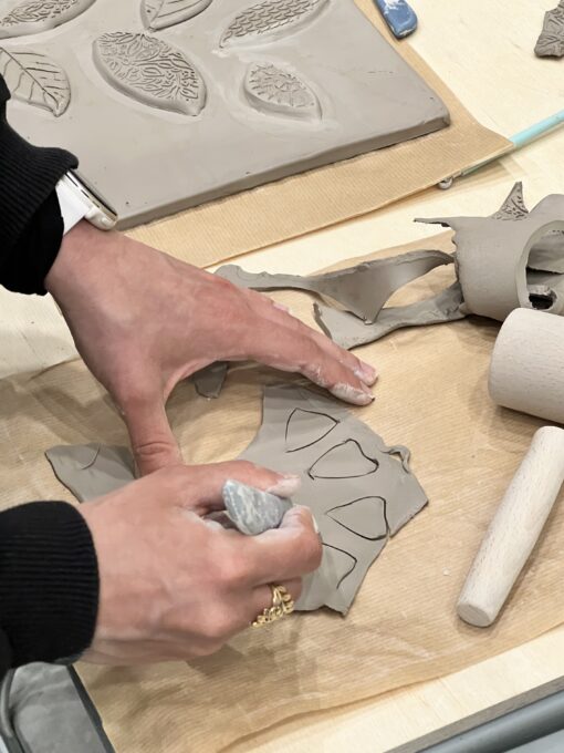 zdjęcie z warsztatów ceramicznych, na pierwszym planie dłonie osoby, która wycina geometryczne kształty w kawałku gliny